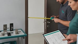מנעולן מודד דלת עם מטר בזמן שלקוחה מציגה תוכנית בטאבלט, ולצידם דגמי מנעולים וידיות